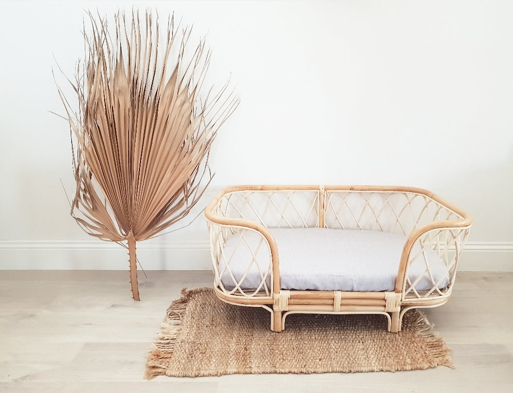 Bella rattan dog bed with a grey sheet sitting in front of a white wall styled with a rattan plant stand and rattan mat. 