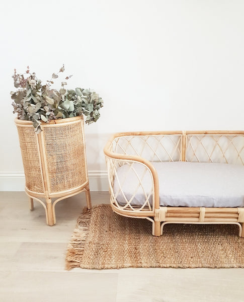 Bella rattan dog bed with a grey sheet sitting in front of a white wall styled with a rattan plant stand and rattan mat. 